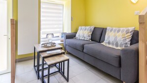 A compact living space with a sofa, two matching pillows, a small table with a tray, and a window with blinds. - Charming Cottage near River & Park, 10km from Nantes, Free Parking, WiFi, Baby-Friendly (Vertou)