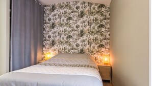 A bedroom with a bed, two bedside lamps, a curtain, and a wall with a tropical wallpaper. - Charming Cottage near River & Park, 10km from Nantes, Free Parking, WiFi, Baby-Friendly (Vertou)