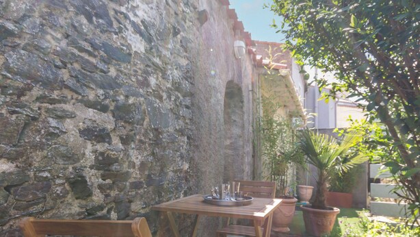 An outdoor seating area with a stone wall, wooden chairs, and a table set for two. - Charming Cottage near River & Park, 10km from Nantes, Free Parking, WiFi, Baby-Friendly (Vertou)