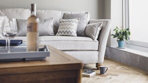A living room with a grey sofa, patterned cushions, a wooden coffee table, a potted plant, and a window with a view. - Oceanwalk - sleeps 8 guests  in 4 bedrooms (Perranporth)