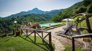 Zona de piscina con sillas de descanso y sombrillas, rodeada de exuberante vegetación y montañas al fondo.