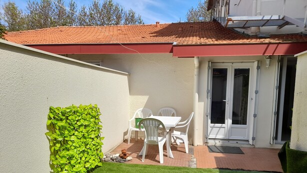 Un patio con césped artificial, una mesa y sillas blancas, un muro cubierto de plantas verdes y un techo rojo.