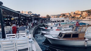 Ein Restaurant am Wasserufer mit Terrassenplätzen, in der Nähe liegen Boote und im Hintergrund ist die Stadt zu sehen.