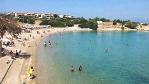 Ein Strand mit klarem Wasser, Menschen beim Schwimmen und Sonnenbaden, und Gebäuden im Hintergrund.