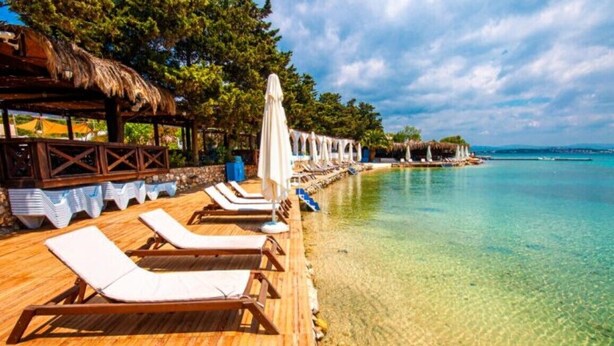 Eine Holzterrasse mit Liegestühlen und Sonnenschirmen, ein Gebäude mit Strohdach und ein klares, türkisblaues Meer.