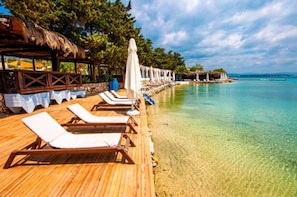 A wooden deck with sun loungers and umbrellas, a thatched-roof structure, and a clear turquoise sea.
