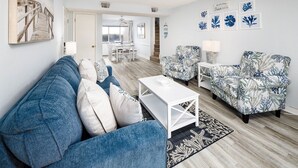 Ein Wohnzimmer mit einem blauen Sofa, einem weißen Couchtisch und gemusterten Sesseln. Im Hintergrund ist ein Essbereich mit Tisch und Stühlen zu sehen.