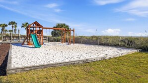 A playground with a swing set and a slide on a sandy surface, surrounded by a wooden fence and palm trees.