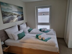 A hotel room with a bed, pillows, a blanket, a remote control, and a painting of a beach scene.