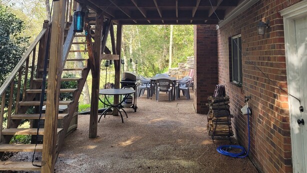 A covered outdoor deck with a table and chairs, a wooden staircase, and a brick house in the background. - Riverside Getaway - Enjoy Quiet Life Right on the Duck River (Shelbyville)