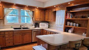 A kitchen with wooden cabinets, a white countertop, a stainless steel dishwasher, a microwave, and a bar area with stools. - Riverside Getaway - Enjoy Quiet Life Right on the Duck River (Shelbyville)