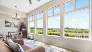 A spacious living room with large windows, a chandelier, a dining area, and a view of greenery and a distant building. - Beautiful Walk-in Lake View Condo with HA Features • Sunroom + Covered Patio (Branson)
