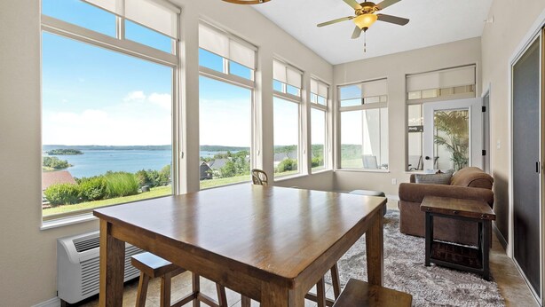 A spacious room with a large wooden table, a ceiling fan, and a view of a body of water. - Beautiful Walk-in Lake View Condo with HA Features • Sunroom + Covered Patio (Branson)