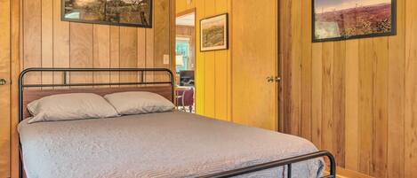 A wooden-paneled room with a bed, two framed pictures, and a door leading to another room.