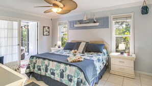 Quarto com cama grande, mesa de cabeceira, ventilador de teto e janela com vista para o verde.