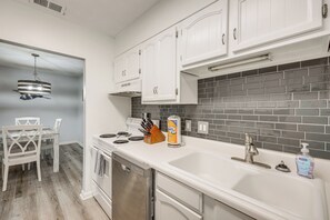 A kitchen with white cabinets, a stainless steel dishwasher, a white sink, and a grey tiled backsplash.