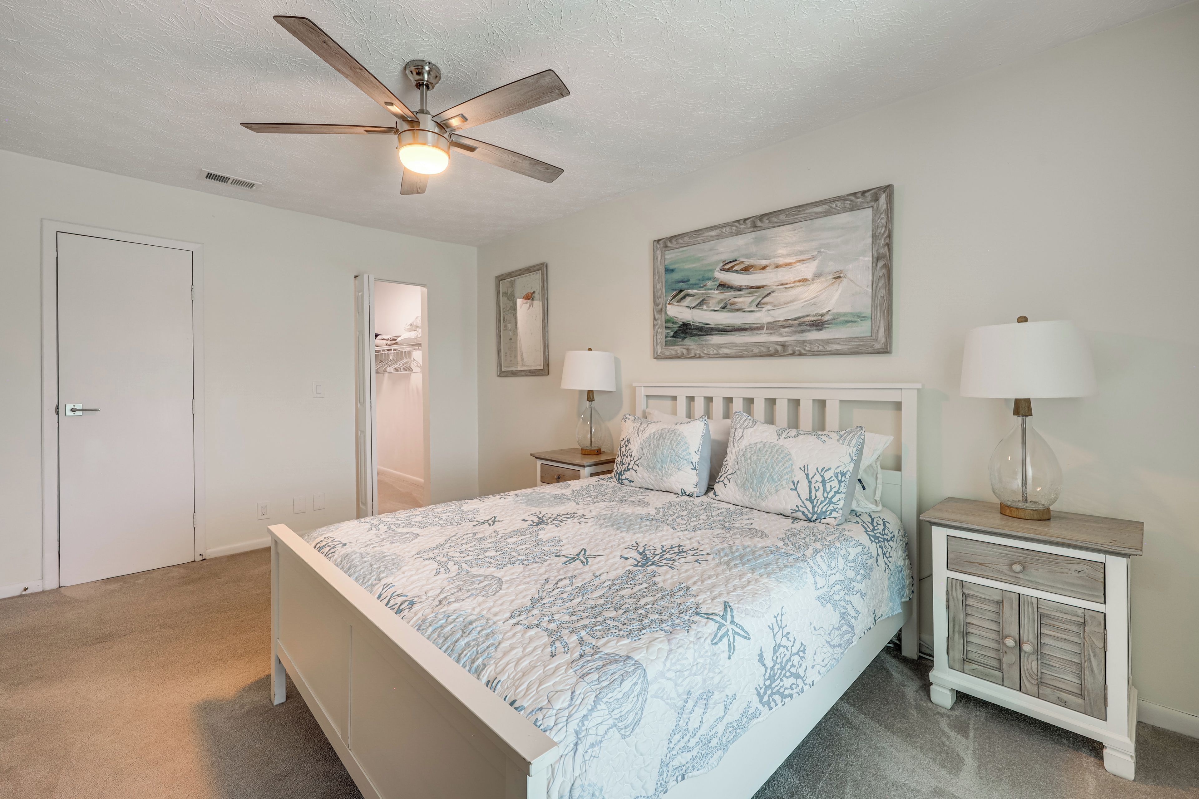 A bedroom with a bed, a ceiling fan, a nightstand with a lamp, a painting on the wall, and a door.