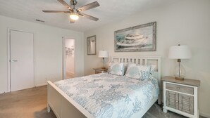 A bedroom with a bed, a ceiling fan, a nightstand with a lamp, a painting on the wall, and a door. - < Half-Mi to Fort Clinch Beach! Condo w/ Balcony (Fernandina Beach)