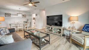 A living room with a flat-screen TV, a ceiling fan, a dining area with chairs and a table, and a sofa. - < Half-Mi to Fort Clinch Beach! Condo w/ Balcony (Fernandina Beach)