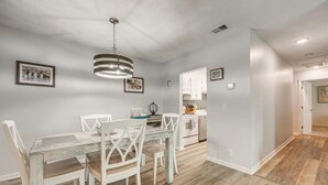 A dining area with a wooden table, white chairs, a hanging light fixture, and a kitchen in the background. - < Half-Mi to Fort Clinch Beach! Condo w/ Balcony (Fernandina Beach)