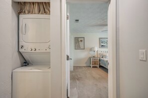 Ein Waschraum mit Waschmaschine und Trockner, ein Gang, der zu einem Schlafzimmer mit Bett und Nachttisch führt.