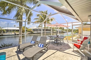 A covered outdoor area with lounge chairs, a table, and a view of a water body and buildings in the background.
