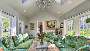 A spacious living room with a high ceiling, large windows, and a chandelier. There are multiple seating areas with colorful patterned upholstery and a central table with a vase of flowers. - Pittsford ‘Trolley Home’ - Near Erie Canal! (Pittsford)