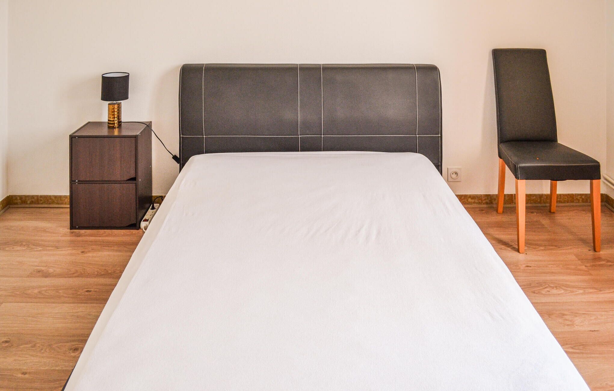A bed with a white sheet, a dark headboard, a nightstand with a lamp, and a chair.