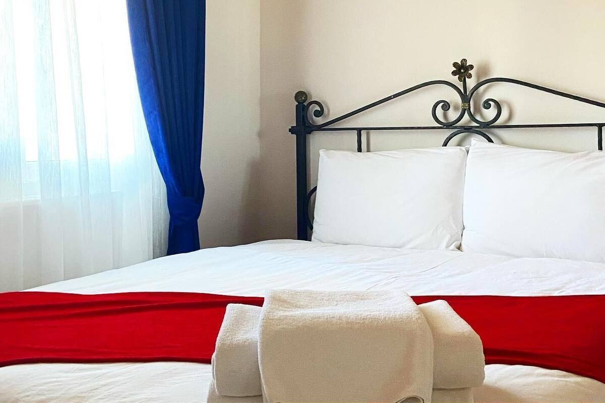 A neatly made bed with white linens and a red blanket, a metal headboard with floral details, and a window with blue curtains.