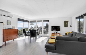 A modern living room with a grey sofa, wooden cabinet, and a dining area with chairs and a table.