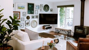 A living room with a white sofa, a bench, a flat-screen TV, a fireplace, and various framed pictures on the wall. - The Grange country retreat, with a birds eye view! (Mittagong)
