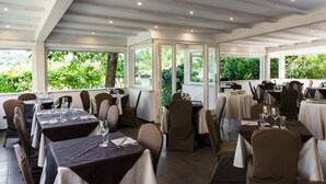 Un restaurant avec des tables dressées pour le service, de grandes fenêtres et une vue sur la verdure environnante.