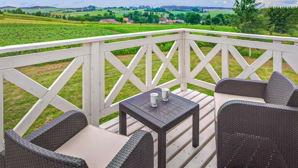 A covered outdoor seating area with wicker furniture and a table, overlooking a green landscape with fields and houses. - 3 bedroom amazing home in Sterlawki Male (Sterlawki Male)
