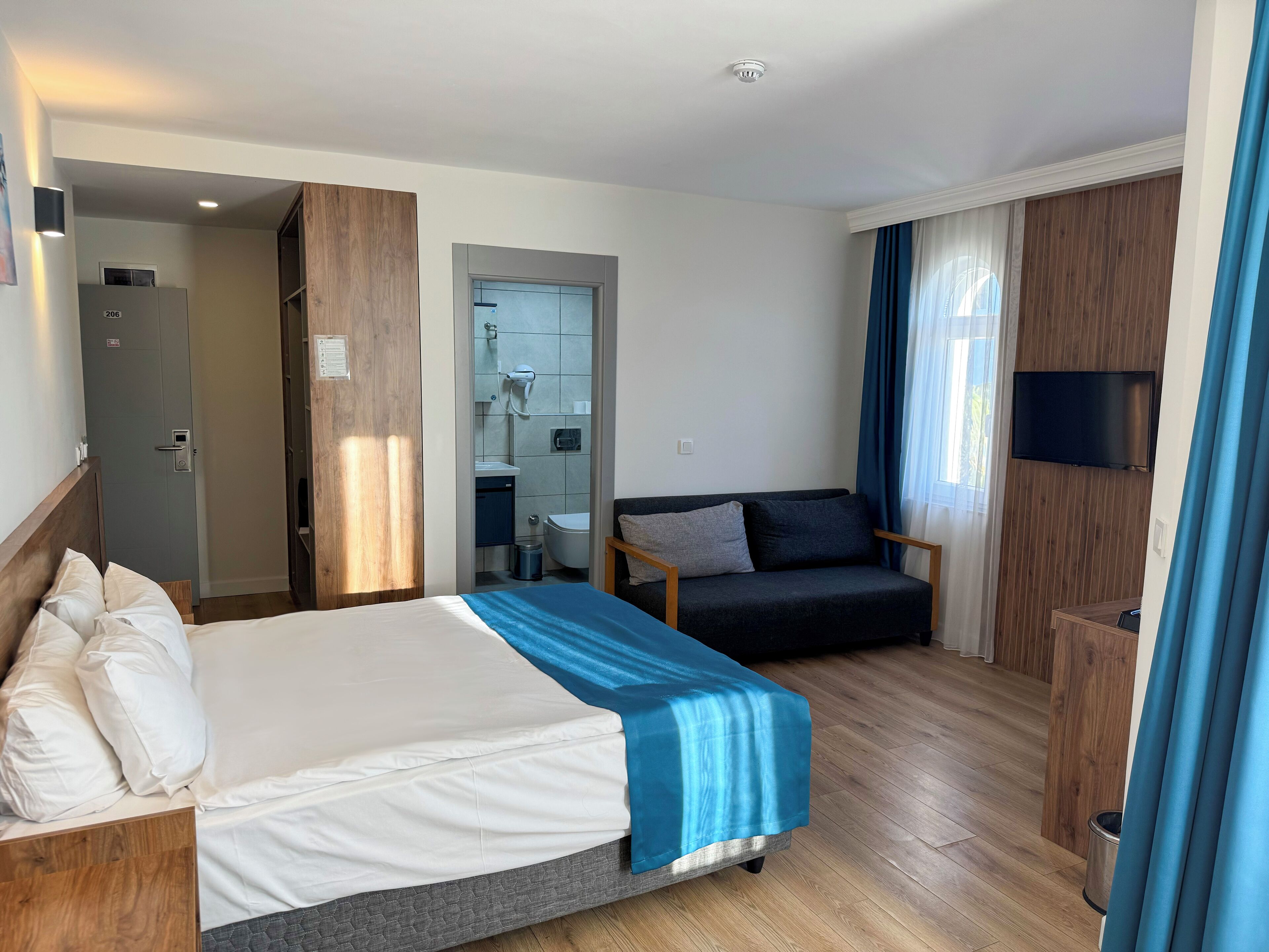 Habitación de hotel con cama, sofá, televisión y baño visible a través de una puerta abierta.