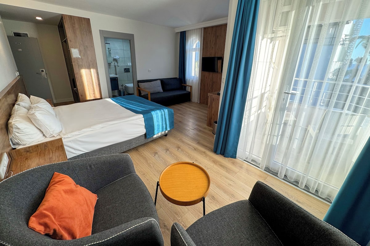 A hotel room with a bed, two armchairs, a small table, a sofa, a TV, and a bathroom visible through an open door.