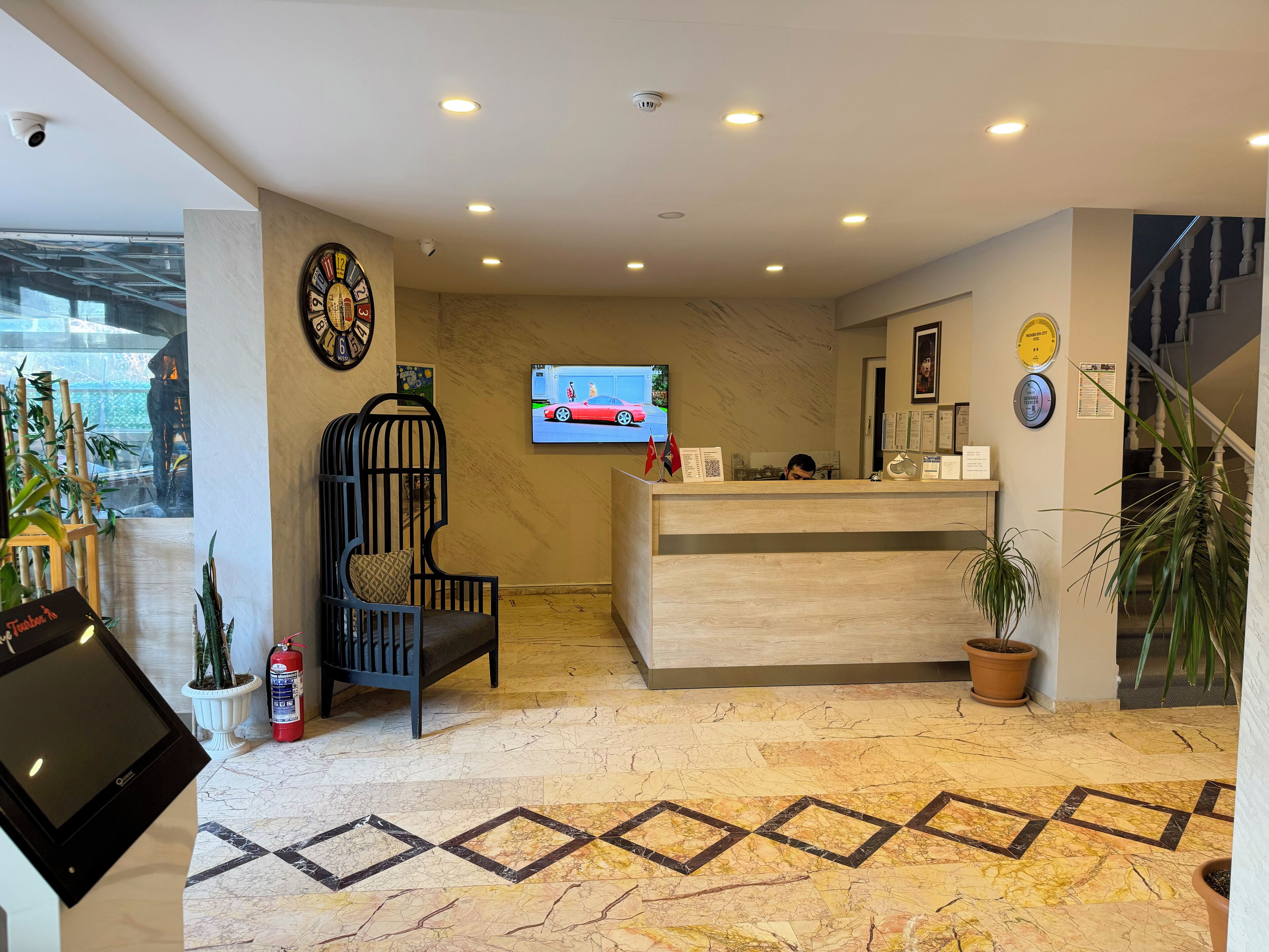 Zona de recepción de un hotel con un mostrador de madera, un reloj, un televisor mostrando un automóvil rojo y un extintor.