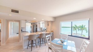 Een moderne keuken met witte kasten, een centraal kookeiland met barstoelen, en uitzicht op het water door grote ramen.