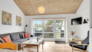 Un salon moderne avec un canapé, une table basse et des fauteuils, un plafond en bois et une grande fenêtre donnant sur l’extérieur.