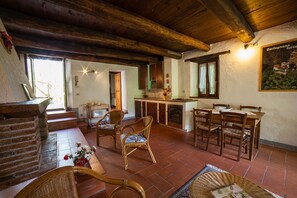 Un intérieur rustique avec une cheminée en pierre, une table à manger en bois, des chaises en osier et un coin cuisine doté d’un comptoir et de placards en bois.