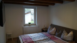 A bedroom with a bed, a window, a radiator, a small stool, and a nightstand with a lamp. - Vacation home "Lisbeth" (Edesheim)