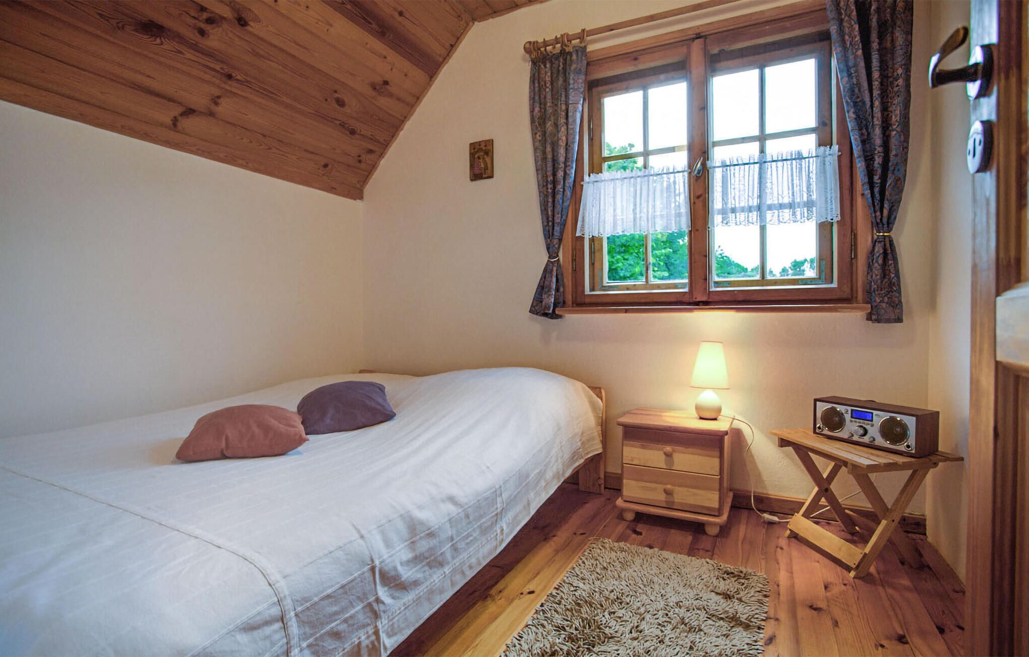 Ein Schlafzimmer mit einem Bett, einem Nachttisch mit Lampe, einem Radio und einem Fenster mit Vorhängen.