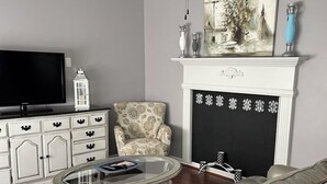 A living room with a sofa, two armchairs, a glass coffee table, a TV, a fireplace, and a ceiling fan.