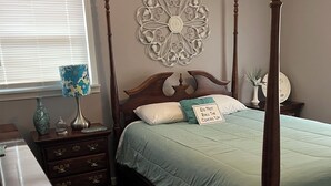 A bedroom with a four-poster bed, a decorative wall piece, a bedside table with a lamp, and a window with blinds.