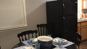 A dining area with a round table set for four, surrounded by chairs. A black refrigerator is in the background, and a window with blinds is on the left.
