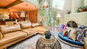 Un salon chaleureux avec un canapé en cuir, un canapé bleu et une table basse en rotin. Un escalier mène à un niveau supérieur, et diverses plantes décorent l’espace.