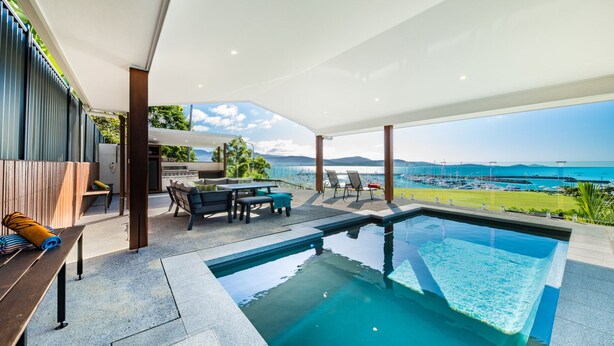 Un espace piscine extérieur moderne avec vue sur la mer, un coin barbecue et des sièges sous une terrasse couverte.