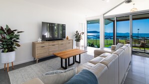 Un salon moderne doté d’un grand téléviseur, d’une table basse en bois et d’un canapé. De grandes portes vitrées offrent une vue sur une marina et des montagnes.