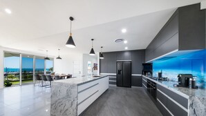 Une cuisine moderne dotée d’un grand îlot central, de meubles de rangement aux couleurs sombres et offrant une vue sur l’océan.