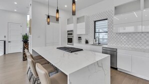 Una cocina moderna con armarios blancos, una isla grande, iluminación colgante y vistas al exterior.