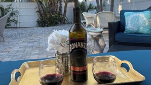 Una botella de vino Bonanza, dos copas de vino tinto y una flor blanca sobre una bandeja en un entorno al aire libre con palmeras y un mantel azul.